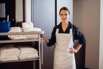 A smiling hotel maid standing with a cleaning cart in a corridor.