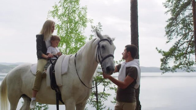 Tracking shot of blond woman and cute little boy sitting on white horse as bearded man petting it. Lake with scenic view in background