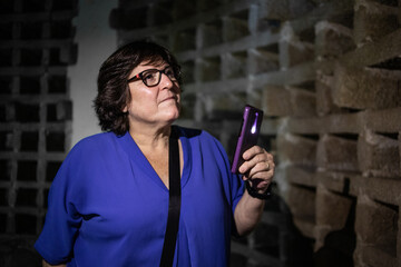 Fototapeta premium retrato de señora dentro de un antiguo palomar, para poder ver alumbra con la linterna del movil. Lleva gafas y una camisa azul. Galicia, España
