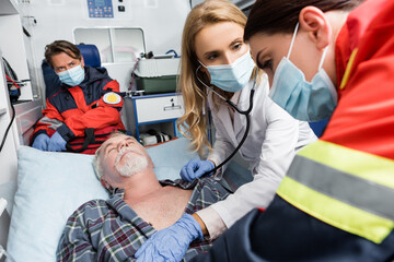 Selective focus of doctor with stethoscope looking at paramedic in medical mask and sick patient in...