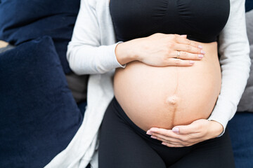 Portrait of young pregnant female sitting on blue sofa at home. Caucasian pregnant woman in black sports outfit touching her belly.