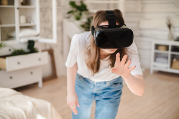 Young woman in jeans and white T-shirt wearing virtual reality helmet plays game, works, watches video in room next to bed. Concept of modern technology, VR, augmented virtual games, entertainment.