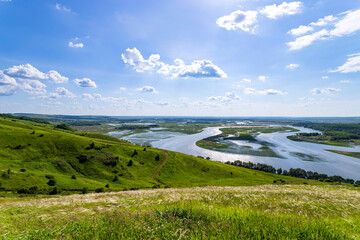 Fototapeta na wymiar Sunny summer landscape with river,fields,green hills and beautiful clouds in blue sky.Pretty view of summer scenery on a nice day.Green trees growing along the river.