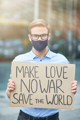 Vertical of young male activist wearing black protective mask holding banner or poster and looking at camera while protesting against the war outdoors, standing on the road