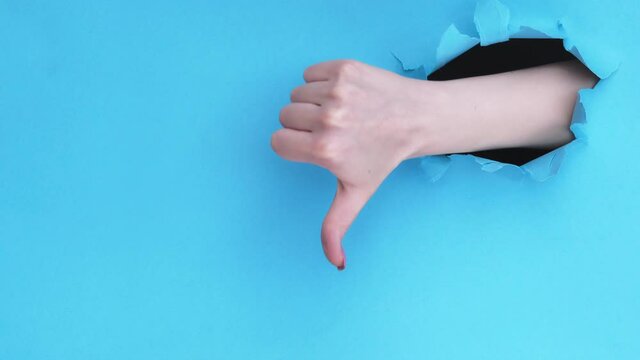 Dislike gesture. Wrong choice. Female hand showing thumb down inside breakthrough paper hole isolated on blue ripped wall background with copy space. Rejection sign set of 2 loop. Not cool. Bad idea.