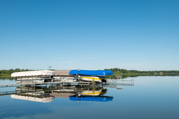 Fototapeta na wymiar Docked boats on lifts are reflected in the calm water of a pretty lake in Minnesota, on a sunny summer day with beautiful clear blue sky.