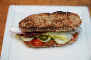 Sandwich of toasted farmer's bread with tomato, cucumber slices, jalapeno slices, lettuce and radicchio on a white square plate on a rustic wooden table
