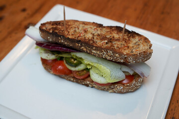 Sandwich of toasted farmer's bread with tomato, cucumber slices, jalapeno slices, lettuce and radicchio on a white square plate on a rustic wooden table