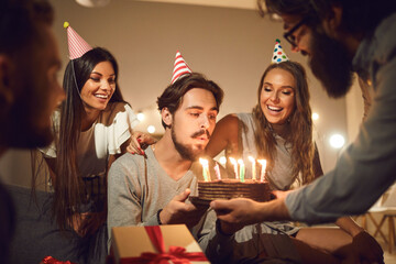 Young man blowing candles on his birthday cake surrounded by happy smiling friends