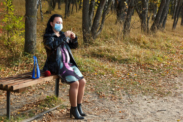 pregnant brunette woman on a park bench in a medical mask knits for the unborn child.