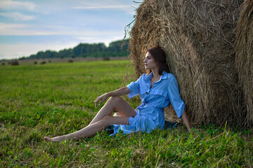 Naklejka na ściany i meble A girl in a blue dress near a haystack on a green field with a blue sky