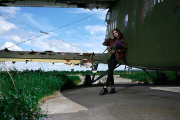 a girl in a brown leather jacket and black jeans with a bouquet of lupins stands near an old broken plane