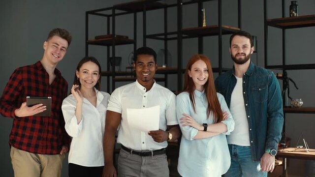 Portrait of cheerful diverse business team standing together in office, looking at camera. Wide shot of multinational young business people or company staff posing in conference room.