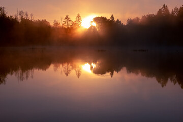 Landscape sunrise on the lake in lilac fog. The calm surface of the water and the reflection of the sun's rays