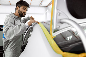 caucasian bearded auto mechanic in uniform stick the tape on car body, going to polish and repair it