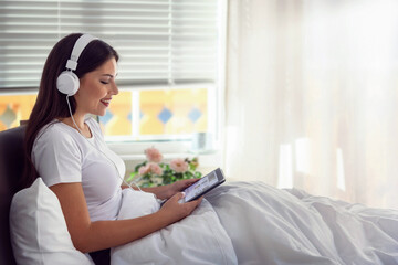 A beautiful, young woman wearing headphones and reading the morning news on her tablet device in bed wilst listening to music