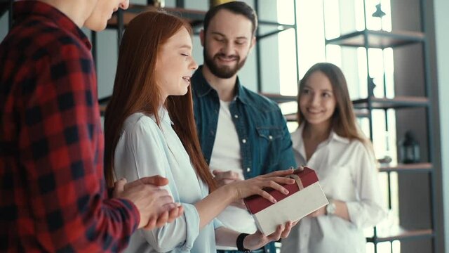 Side view of cheerful attractive young woman opening present given by colleagues in office to applause of coworkers. Happy team of young business people congratulate woman colleague and give gift.