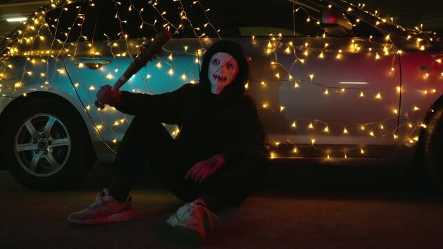 A man in a bloody mask sits on the pavement. The killer is ready for the Purge. A baseball bat wrapped in barbed wire for Halloween. The dangerous car is decorated with a garland.