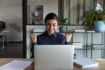 Excited millennial indian female employee look at laptop screen feel euphoric read promotion news online. Happy overjoyed young ethnic woman triumph get good pleasant message on computer in office.