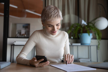 Smiling young female employee sit at desk work with paperwork document using modern smartphone in office. Happy Caucasian woman worker hold cellphone busy considering reading contract at workplace.