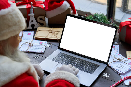 Over shoulder view of Santa Claus wearing costume using laptop computer with white blank empty mock up screen monitor sitting at workshop table on Merry Christmas eve. E commerce website ads concept