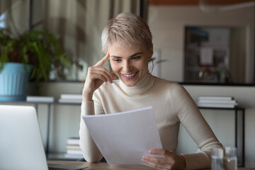Smiling young Caucasian businesswoman sit at desk in office work on computer read post paper...