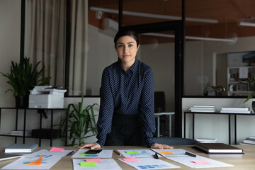 Portrait of serious young indian businesswoman stand pose look at camera at desk in office. Concentrated confident successful 30s mixed race ethnicity female employee show leadership at workplace.