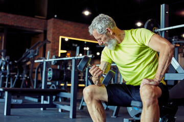 Gaining muscles after 40. Strong mature man in sportswear lifting heavy dumbbells and pumping his...