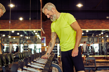 Side view of a handsome mature man in sportswear taking a dumbbells while working out at gym, exercising with weights