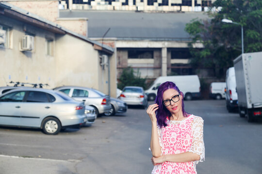 Young Caucasian woman with purple hair and glasses, looking at the camera. Blurred industrial neighborhood background