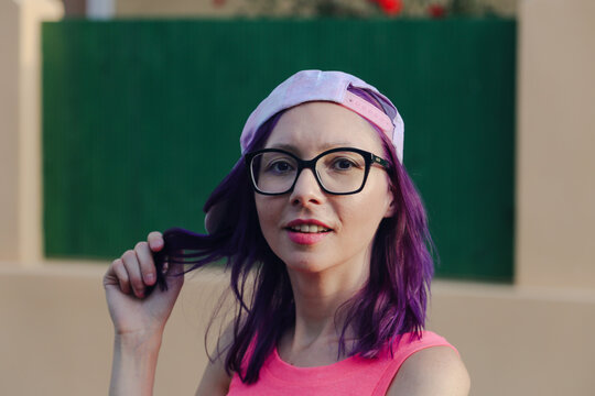Young purple haired Caucasian Woman looking at the camera and playing with her hair, wearing pink cap and shirt
