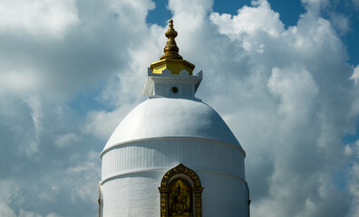 The upper part of the rounded Buddhist Peace Stupa, with a golden spire, against a background of white clouds