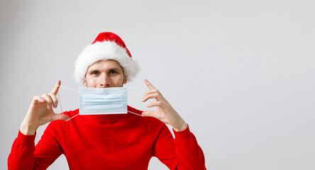Beautiful man wearing disposable protective face mask and Santa hat. Male model wearing red sweater on white isolated. Christmas concept during pandemic of COVID-19 Coronavirus respiratory viruses 