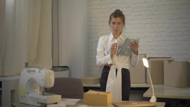 Positive confident Caucasian woman checking online order on tablet and approaching to the table with packed ready products. Portrait of tailor selling clothes on Internet site. Small business concept.