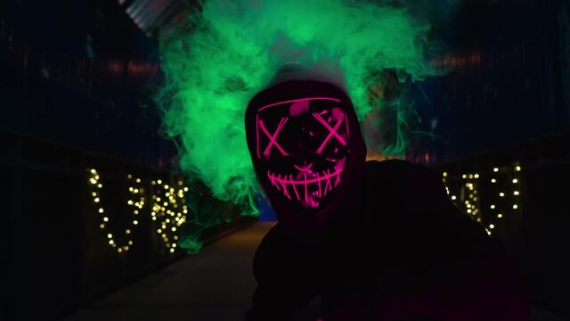 The man in the neon mask blows smoke. Thick green and pink smoke. Scary Halloween Purge killer costume.