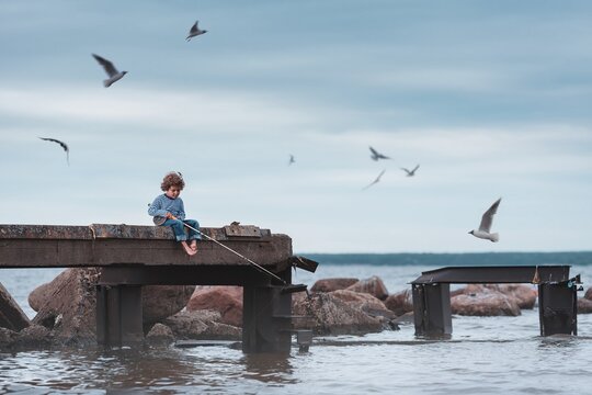 Cute curly-haired kid fishing from a concrete pier on a vintage fishing rod. Gulls are flying around. Haze by the water. Summer evening, overcast. - Powered by Adobe