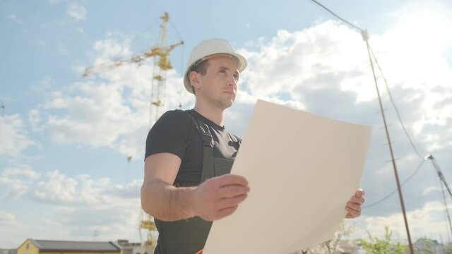 A man in a helmet is holding a construction plan in his hands. A builder is checking blueprints.