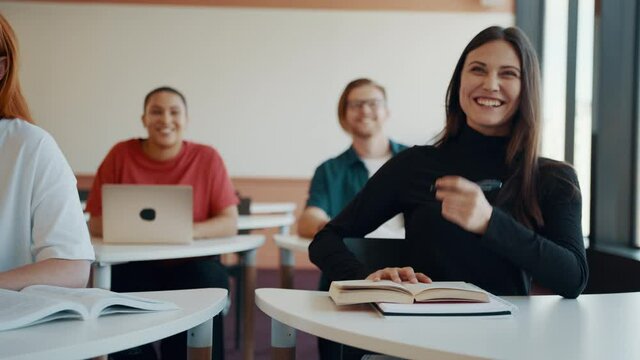 Group of male and female college students sitting in a classroom and smiling. Woman talking to the teacher as she and the class share a laugh during the lecture.
