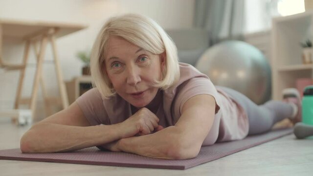 POV portrait with fading of a white-haired sports person lying down on yoga mat on floor of living room facing forward and talking and smiling on camera