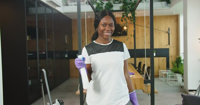 Close up of joyful african american woman holding cleaner spray with hands in gloves and looking in camera smiling. Concept of housekeeping, family cleaning house, lifestyle.