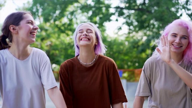 Happy teenage girls walking outdoors in green skate park. Friends having fun, laughing, holding hands. Pretty hipster women with dyed hairstyle in casual wear.