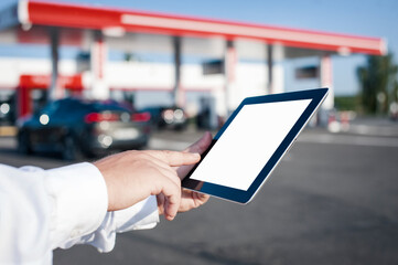 A man holds a close-up of a tablet with a white screen in his hands against the backdrop of a car and a gas station. Technology mockup for apps and websites.