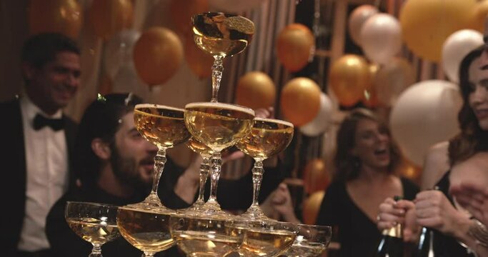 Beautiful young woman dancing around a pyramid of champagne glasses with friends at a party. Tower of champagne glasses with people dancing at new years eve.
