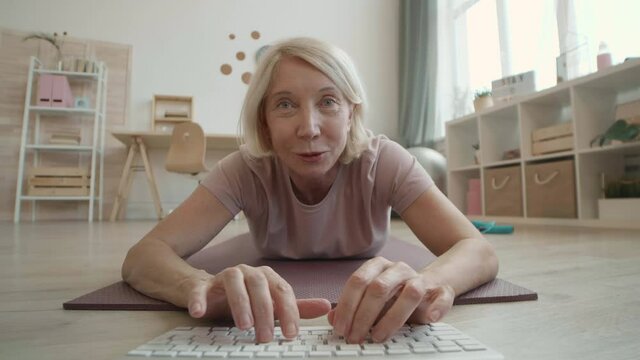 POV of white-haired strong senior female client laying down on yoga mat typing on keyboard talking and smiling on camera to fitness teacher then doing push-ups at home