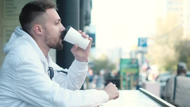 Handsome young hipster student in white jacket is sitting at table in street cafe alone and enjoying drinking hot aromatic coffee from paper Cup. Walk through city streets