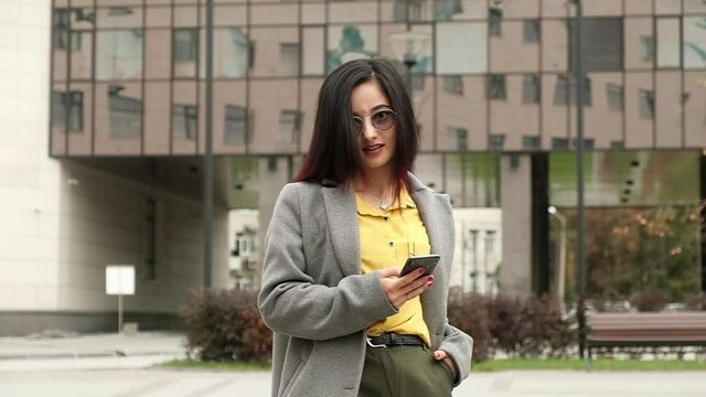 Beautiful brunette with glasses and stylish casual clothes stands on city street and holds phone in hands. stylish young businesswoman uses modern phone on street to communicate and work in apps