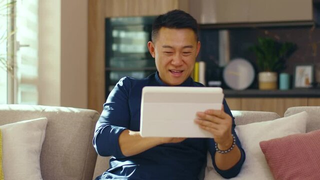 Smiling asian young man use tablet computer sitting on sofa at home. Feel happy. Chinese businessman device handsome. Communication. Close up. Slow motion