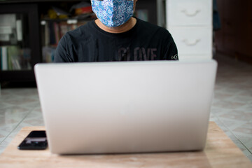 The masked man sat on the floor and worked at home using a laptop and holding a smartphone on a wooden table during the coronavirus outbreak, or COVID-19, taking a close-up, blurred background
