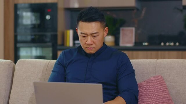 Portrait sad serious asian man sitting on sofa use laptop computer work at home. Network work unhappy guy interior technology social media. Slow motion