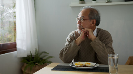 Sad old retired gray haired grandpa asian man sitting alone at table desk at window boring stay...